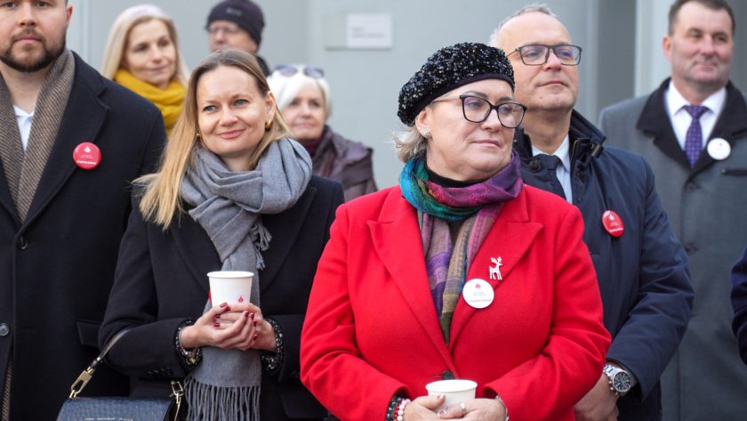 Grupa uczestników uroczystości stoi na zewnątrz, słuchając przemówień. Na pierwszym planie dwie kobiety: jedna w czarnym płaszczu i szarym szaliku trzyma papierowy kubek, druga w czerwonym płaszczu i czarnej ozdobnej czapce również trzyma kubek. W tle stoją inni uczestnicy, elegancko ubrani, część z przypiętymi okolicznościowymi znaczkami