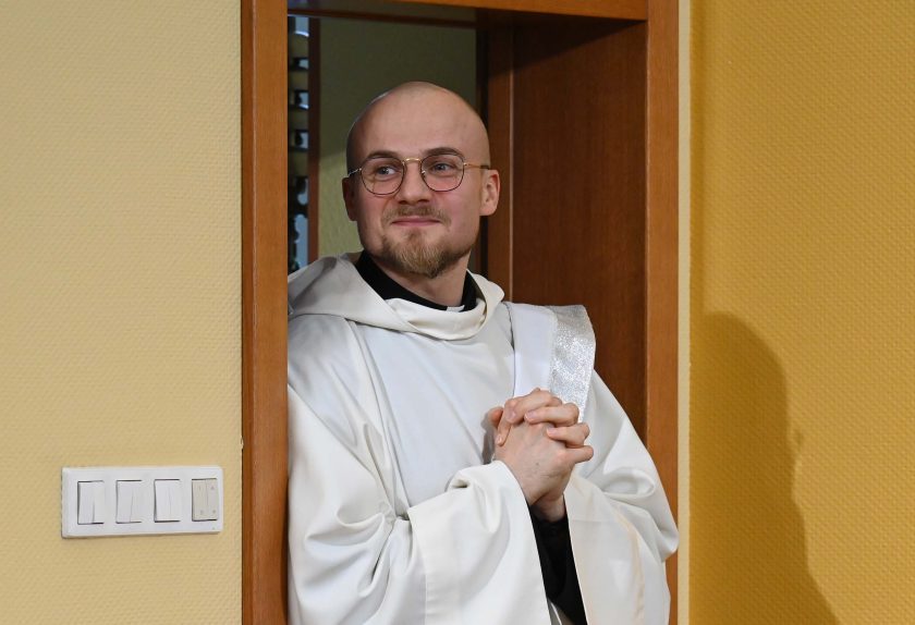 Młody zakonnik w białym habicie stoi w drzwiach kaplicy, z dłońmi splecionymi przed sobą. Uśmiecha się lekko, a za nim widoczna jest jasna ściana i elementy wnętrza zakonnego