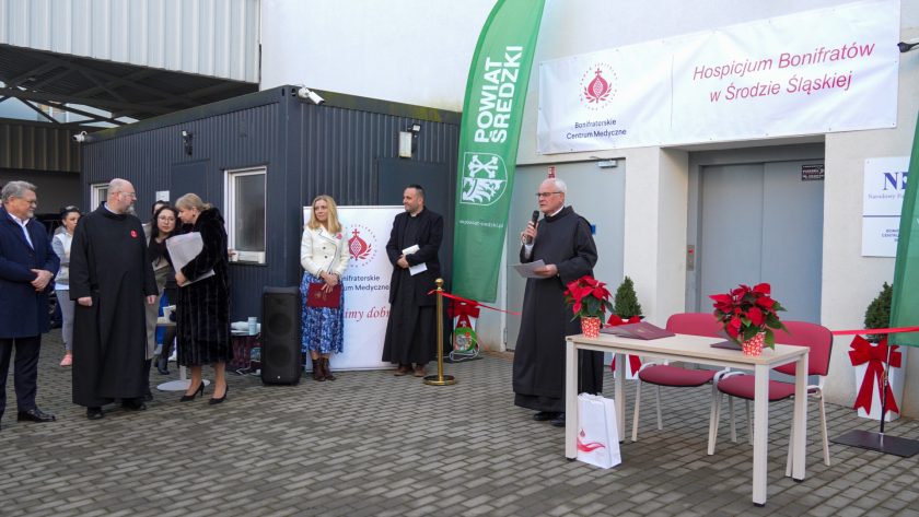 Uroczystość otwarcia hospicjum Bonifratrów w Środzie Śląskiej odbywa się na dziedzińcu przed budynkiem. Po prawej stronie stoi brat zakonny w czarnym habicie, przemawiający do mikrofonu i trzymający dokumenty. Obok znajduje się stolik z dwoma czerwonymi fotelami i doniczkami z poinsecjami. Za stołem wisi biały baner z nazwą hospicjum. Po lewej stronie widać grupę uczestników, w tym osoby duchowne i świeckie, stojących obok zielonego roll-upu z logo Powiatu Średzkiego