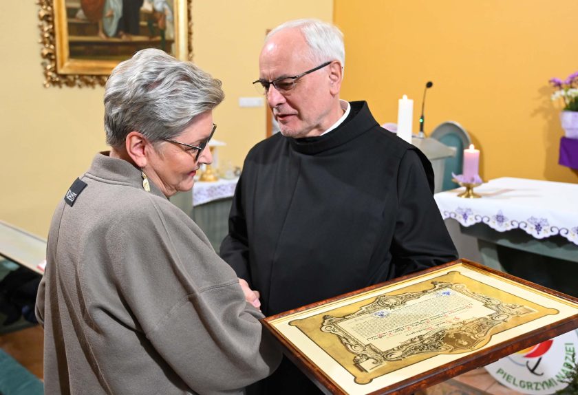 Kobieta o krótkich, siwych włosach odbiera od zakonnika w czarnym habicie oprawiony w ramę dokument. Oboje stoją w pobliżu ołtarza i wymieniają uścisk dłoni. W tle widoczne są świeczniki, stół ołtarzowy i dekoracje kaplicy