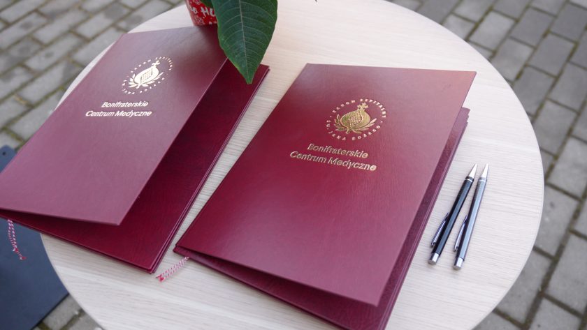 Na okrągłym, jasnym stoliku leżą dwie bordowe, eleganckie teczki z tłoczonym złotym logo Zakonu Bonifratrów i napisem „Bonifraterskie Centrum Medyczne”. Obok znajdują się dwa długopisy. W rogu kadru widoczny jest fragment doniczki z poinsecją