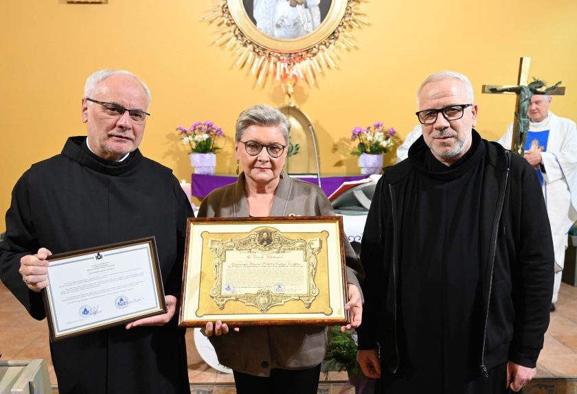 Trzy osoby stoją przed ołtarzem kaplicy: zakonnik w czarnym habicie, kobieta trzymająca duży oprawiony dyplom oraz drugi zakonnik trzymający kolejny dokument w ramie. Za nimi widoczna jest monstrancja i kwiaty na ołtarzu