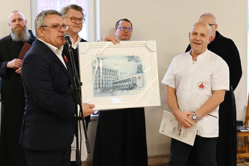 Moment przekazania pamiątkowego obrazu – mężczyzna w garniturze prezentuje oprawioną grafikę budynku (w białej ramie), obok stoi pracownik w białym uniformie z czerwonym logo na kieszeni; w tle widoczni duchowni i goście