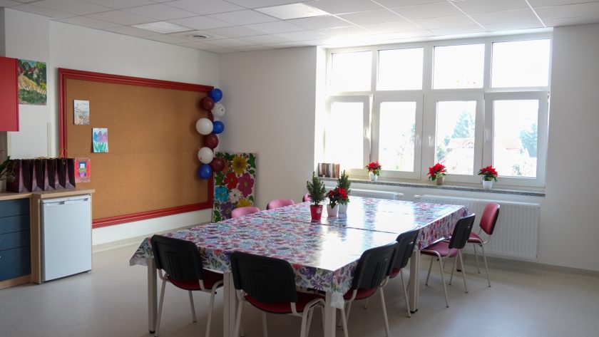 Jasna, przestronna sala z dużym stołem przykrytym kolorowym, kwiatowym obrusem, otoczonym czerwonymi krzesłami. Na parapetach oraz na stole stoją doniczki z poinsecjami i małymi choinkami. W tle widać korkową tablicę, balony dekoracyjne oraz regał z torbami upominkowymi