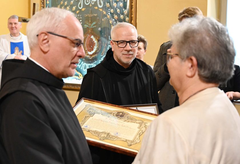 Zakonnik w czarnym habicie przekazuje oprawiony w ramę dokument kobiecie stojącej naprzeciw niego. Obok znajduje się drugi zakonnik, który przygląda się ceremonii. W tle widać obraz i fragment ołtarza