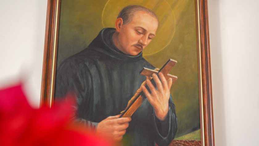 Zbliżenie na obraz przedstawiający św. Jana Bożego w czarnym habicie, trzymającego z czcią krucyfiks. W tle dominuje ciepłe, złotozielone światło, a na pierwszym planie, nieostro, widoczne są czerwone liście poinsecji