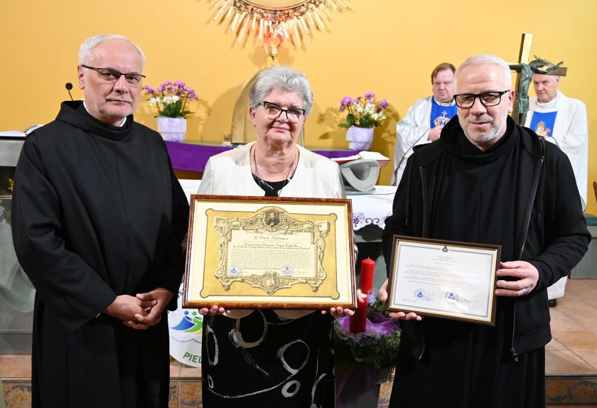 Zakonnik w czarnym habicie, kobieta w okularach trzymająca oprawiony dyplom oraz drugi zakonnik z ramką dokumentu stoją razem przed ołtarzem. Za nimi widoczne są kapłani w białych ornatach, kwiaty i dekoracje liturgiczne