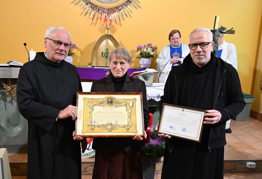Zakonnik i kobieta w czarnym żakiecie stoją z przodu kaplicy, prezentując duży oprawiony dyplom. Obok nich drugi zakonnik trzyma drugi dokument. W tle dwaj kapłani w ornatach oraz ołtarz z kwiatami