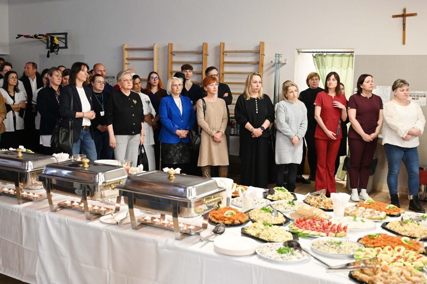 Szeroki plan sali z poczęstunkiem – na pierwszym planie długi stół z podgrzewaczami i półmiskami (sałatki, kanapki, przekąski), za stołem stoi grupa uczestników słuchających przemówienia; na ścianie widać krzyż i sprzęt rehabilitacyjny