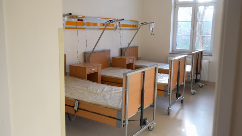 Nowoczesna sala szpitalna z trzema łóżkami wyposażonymi w regulowane barierki i wysięgniki. Przy każdym łóżku znajdują się drewniane szafki. Pomieszczenie jest jasne, oświetlone dużym oknem z widokiem na drzewa