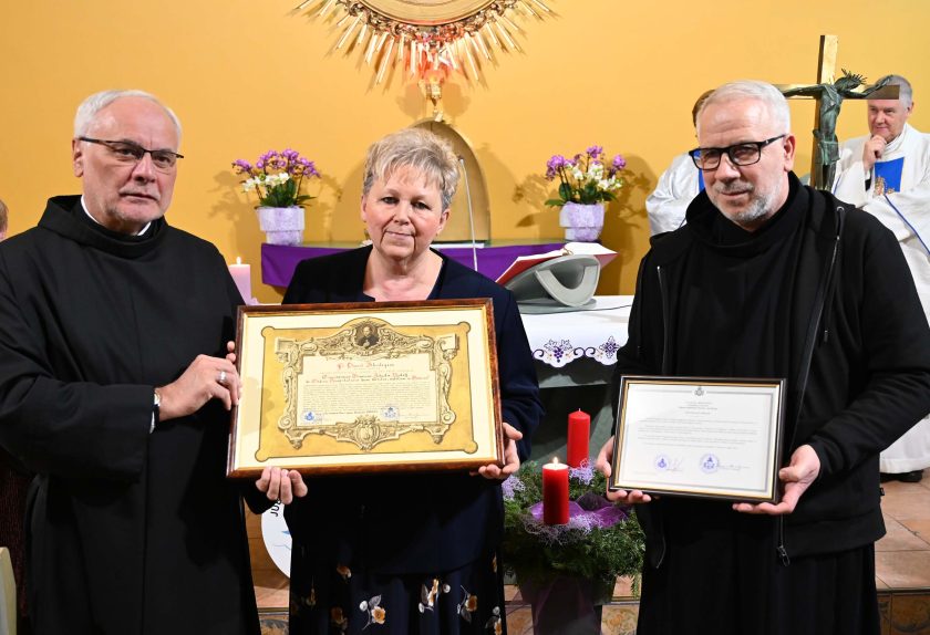Zakonnik, kobieta w granatowym stroju oraz drugi zakonnik prezentują dwa oprawione dyplomy stojąc przed ołtarzem. Za nimi widoczne są elementy liturgiczne oraz kapłani w białych szatach