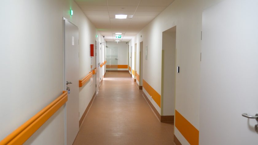 Długi, jasny korytarz w budynku medycznym, utrzymany w biało-pomarańczowej kolorystyce. Na ścianach znajdują się poręcze dla pacjentów, a na suficie rozmieszczone są liczne oświetlenia LED. Na końcu korytarza widoczne są szklane drzwi