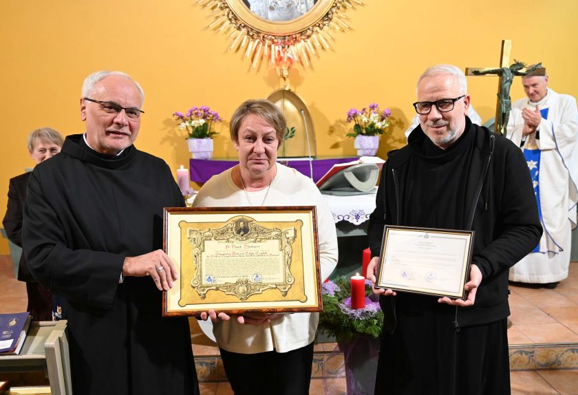 Zakonnik, kobieta w jasnym swetrze trzymająca duży dyplom oraz drugi zakonnik z kolejnym dokumentem stoją przed ołtarzem. W tle widać kapłana w białym ornacie i krzyż