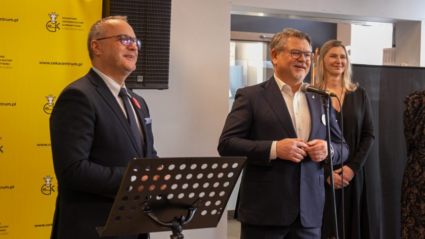 Dwóch elegancko ubranych mężczyzn stoi przed mikrofonami w sali wydarzeń. Jeden mówi do mikrofonu, drugi uśmiecha się, trzymając ręce złożone. W tle widoczne są kobiety uczestniczące w uroczystości oraz żółta ścianka z logo CEKA