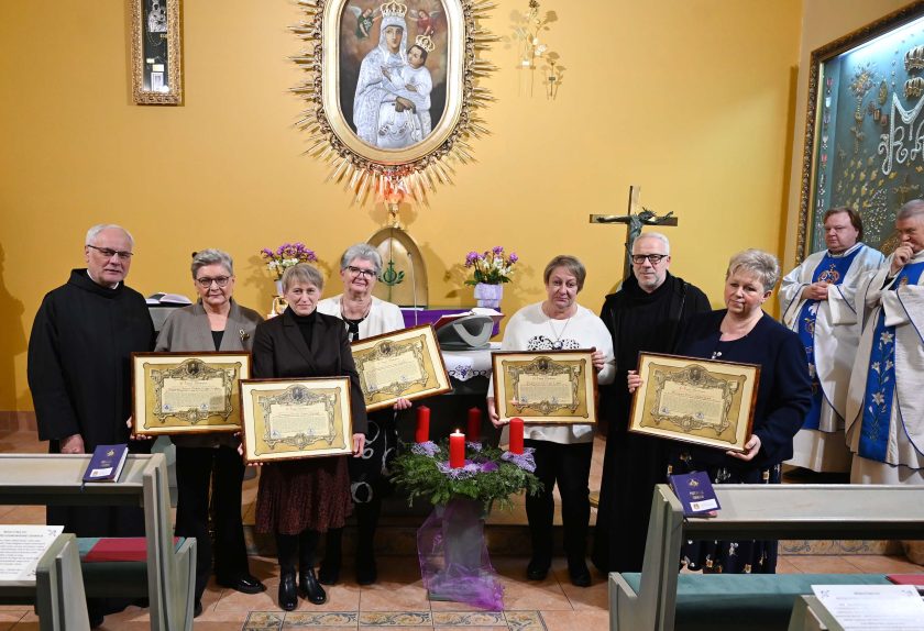 Siedmioosobowa grupa stoi przed ołtarzem kaplicy, każda z kobiet trzyma duży oprawiony dyplom. Po bokach stoją dwaj zakonnicy. Za nimi widoczny jest obraz Maryi z Dzieciątkiem, ołtarz i kapłani w białych ornatach