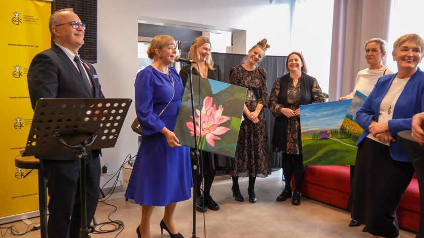 Grupa kobiet stoi na scenie podczas uroczystości. Kobieta w niebieskiej sukience trzyma duży obraz przedstawiający różowy kwiat lotosu, natomiast inna osoba trzyma pejzaż w żywych kolorach. Pozostali uczestnicy uśmiechają się i patrzą w stronę fotografa. Obok nich stoją prowadzący wydarzenie