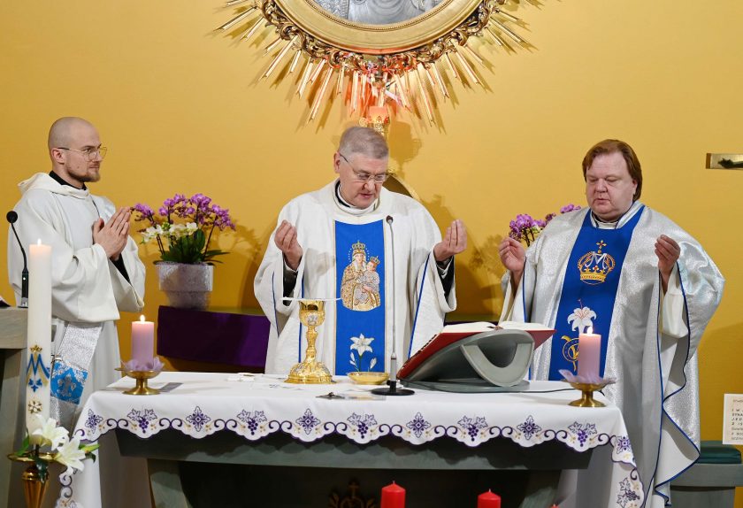 Trzech kapłanów w białych ornatach stoi przy ołtarzu podczas modlitwy eucharystycznej. Kapłan w środku trzyma dłonie uniesione w geście modlitwy. Na ołtarzu znajdują się świece, kielich, księga liturgiczna i dekoracje kwiatowe. W tle wisi obraz Maryi z Dzieciątkiem