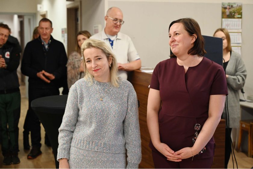 Dwie kobiety stoją obok siebie podczas spotkania – jedna w jasnym, dzianinowym komplecie uśmiecha się do obiektywu, druga w bordowym stroju medycznym patrzy w bok; w tle recepcja i zgromadzeni goście