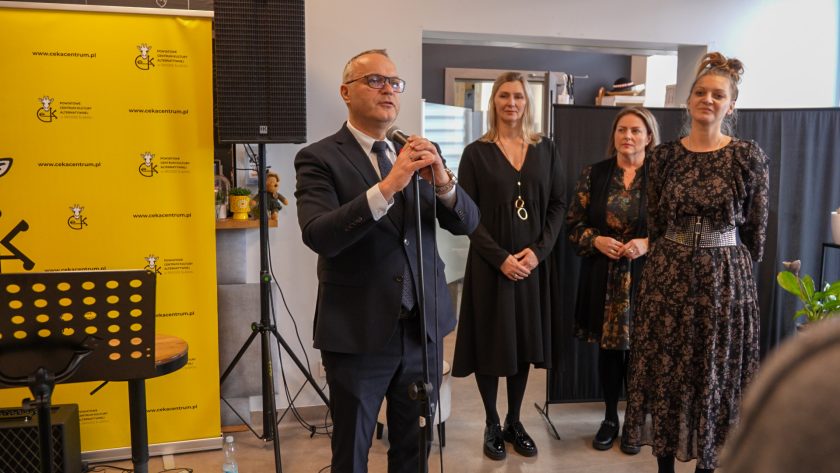 Mężczyzna w garniturze przemawia do mikrofonu na tle żółtej ścianki z logo CEKA. Za nim stoją trzy kobiety ubrane w ciemne stroje, słuchające jego wypowiedzi. Scena odbywa się w nowoczesnym, jasnym wnętrzu