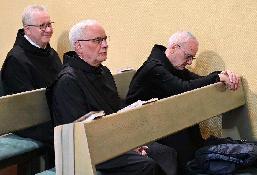 Trzech zakonników w czarnych habitach siedzi w ławce kaplicy. Dwóch z nich patrzy przed siebie, a jeden modli się z pochyloną głową i złożonymi rękami. Na ławce leżą otwarte książeczki liturgiczne