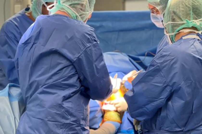 Zespół trzech chirurgów w niebieskich fartuchach i zielonych czepkach przeprowadza zabieg operacyjny na sali operacyjnej, stojąc po obu stronach pacjenta przykrytego sterylnymi obłożeniami