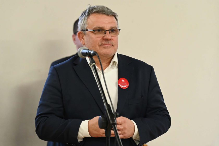 Mężczyzna w granatowym garniturze stoi przy mikrofonie; ma okulary i przypiętą czerwoną przypinkę na klapie marynarki, dłonie trzyma złączone na wysokości pasa, za nim niewyraźne tło sali