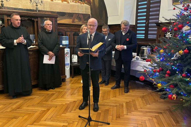 Odczytanie fragmentu Ewangelii podczas spotkania opłatkowego, przy mikrofonie mężczyzna w garniturze trzymający księgę, obok duchowni i przedstawiciele szpitala