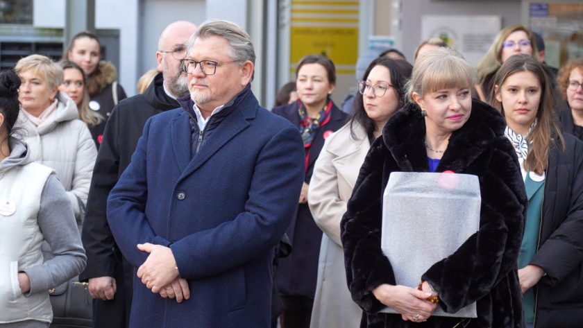 Zebrani uczestnicy stoją blisko siebie, uważnie słuchając. Na pierwszym planie mężczyzna w granatowym płaszczu z rękami splecionymi przed sobą oraz kobieta w czarnym futrzanym płaszczu trzymająca duże zapakowane pudło. Za nimi widoczne są kolejne osoby, w tym zakonnicy oraz pracownicy medyczni