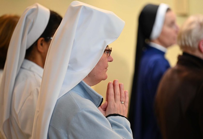 Zbliżenie na siostrę zakonną w białym welonie i jasnej bluzie, stojącą bokiem z dłońmi złożonymi do modlitwy. Obok niej widać inne siostry zakonne, a w tle osoby uczestniczące w nabożeństwie