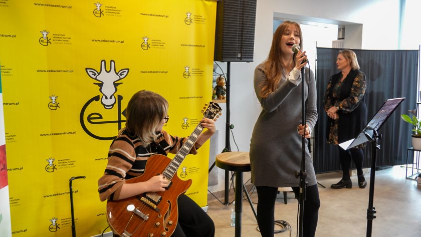 Młoda wokalistka w szarej sukience stoi przy mikrofonie i śpiewa, a obok niej siedzi gitarzystka grająca na elektrycznej gitarze o drewnianym wykończeniu. W tle widoczna jest jaskrawożółta ścianka z logo Powiatowego Centrum Kultury Alternatywnej w Środzie Śląskiej. Z boku stoi kobieta obserwująca występ