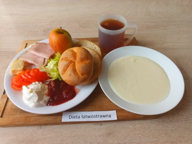 Śniadanie w diecie łatwostrawnej: jasna zupa mleczna, bułka pszenna, pieczywo, wędlina drobiowa, serek twarogowy, pomidor, sałata, jabłko oraz herbata, podane na drewnianej desce