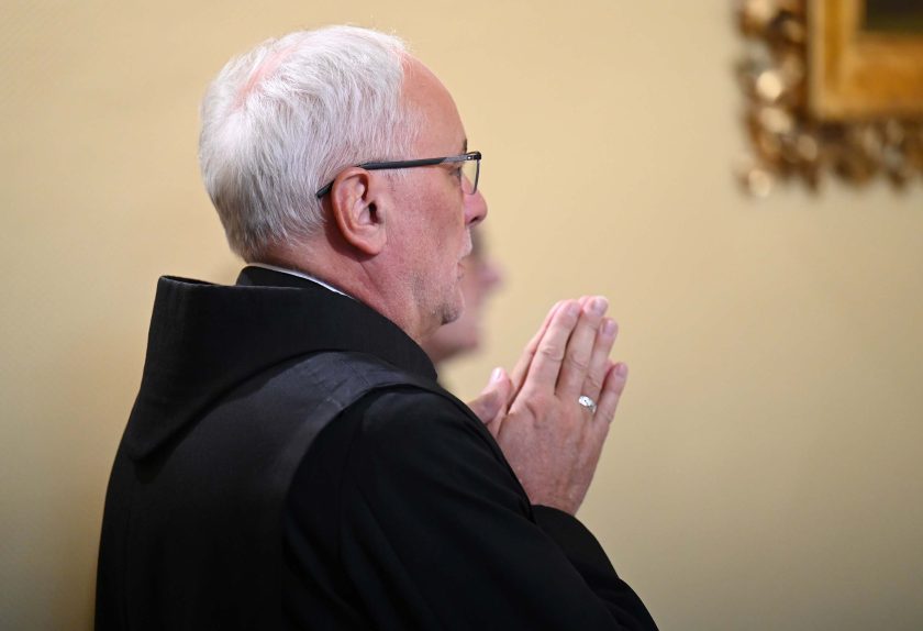 Zakonnik o siwych włosach, widoczny z profilu, stoi z dłońmi złożonymi do modlitwy. Ubrany jest w czarny habit i okulary. W tle rozmyty fragment obrazu w ozdobnej ramie