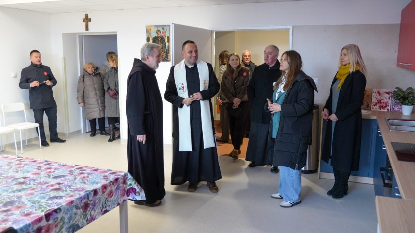 Grupa osób zwiedza wnętrze placówki. W centrum kadru stoi dwóch zakonników – jeden w czarnym habicie, drugi w sutannie i białej stule – rozmawiających uśmiechnięcie. Wokół nich stoją uczestnicy wydarzenia, część w kurtkach zimowych. Na ścianie za nimi wiszą krzyż i obraz religijny. Z lewej strony kadru widoczny jest fragment stołu przykrytego kolorowym obrusem