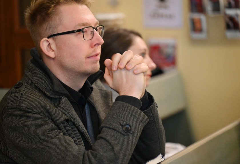 Mężczyzna w okularach i ciemnym płaszczu siedzi w ławce kościoła, trzymając dłonie splecione przy twarzy w geście modlitwy. Patrzy przed siebie ze skupieniem. Za nim siedzi kobieta, również zamyślona