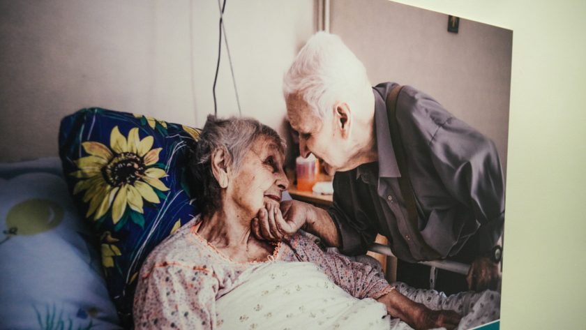 Zbliżenie na dużą fotografię wiszącą na ścianie. Przedstawia ona starszą, leżącą w łóżku kobietę, patrzącą z czułością na pochylającego się nad nią mężczyznę, który delikatnie trzyma jej twarz w dłoni. Oboje mają łagodne, pełne bliskości wyrazy twarzy