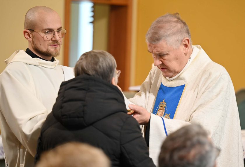 Kapłan w białym ornacie z niebieskim wizerunkiem Maryi udziela Komunii starszej kobiecie w czarnej kurtce. Obok stoi młody zakonnik w białym habicie, obserwując obrzęd. W tle widoczna jest ściana kaplicy