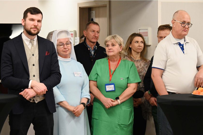 Grupa uczestników uroczystości w korytarzu/sali – mężczyzna w garniturze i kamizelce stoi z przodu, obok zakonnica w jasnym habicie, personel medyczny w zielonym stroju i inni goście; wszyscy skupieni na wydarzeniu poza kadrem