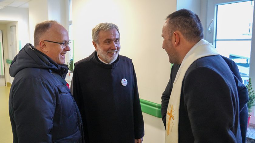 W jasnym szpitalnym korytarzu trzech mężczyzn uśmiecha się podczas rozmowy. Jeden z nich nosi zimową kurtkę, drugi habit zakonny, a trzeci sutannę ze stulą ozdobioną złotym krzyżem. W tle widać okno i świąteczną dekorację roślinną