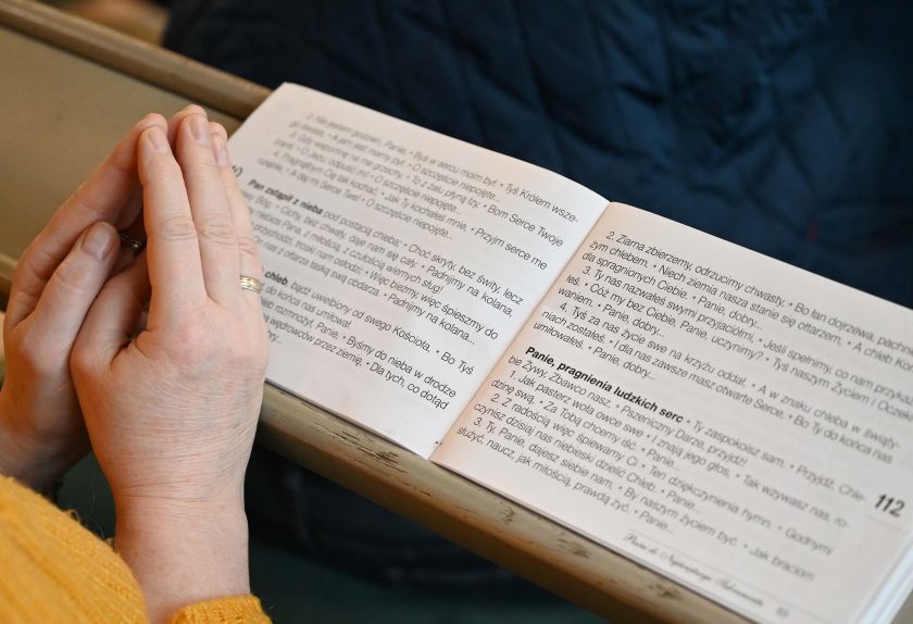 Dłonie osoby trzymającej palce złożone w geście modlitwy spoczywają nad otwartym modlitewnikiem z drukowanym tekstem pieśni i modlitw. Książka leży na ławce, a w tle widać fragment ciemnoniebieskiej kurtki