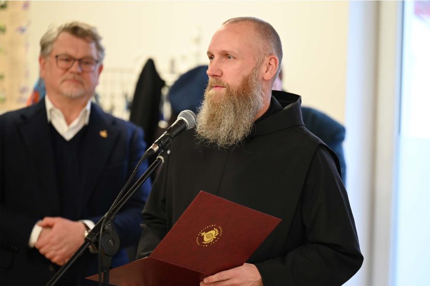 Duchowny w czarnym habicie przemawia do mikrofonu, trzymając bordową teczkę z okrągłym złotym emblematem; w tle, lekko rozmyty, stoi mężczyzna w garniturze