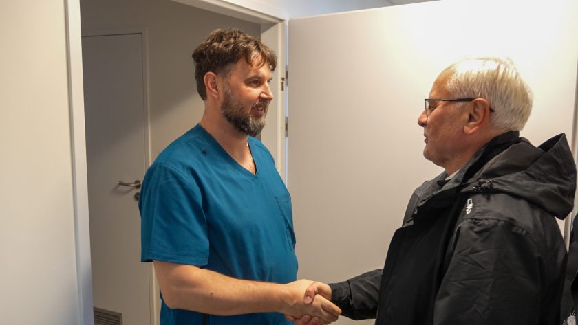 W niewielkim pomieszczeniu mężczyzna w niebieskim uniformie medycznym ściska dłoń starszego mężczyzny w okularach, ubranego w czarną kurtkę. Obaj uśmiechają się uprzejmie. Za nimi znajdują się otwarte drzwi prowadzące do korytarza