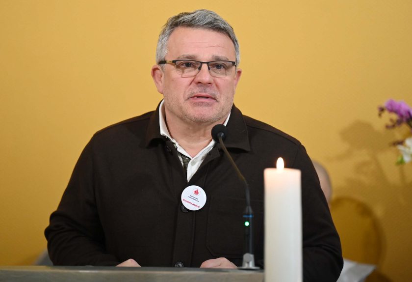 Mężczyzna w okularach i brązowej marynarce mówi do mikrofonu przy ambonie. Na piersi ma przypięty biały znaczek z czerwonym napisem. Przed nim stoi zapalona biała świeca. Tło stanowi żółta ściana kaplicy