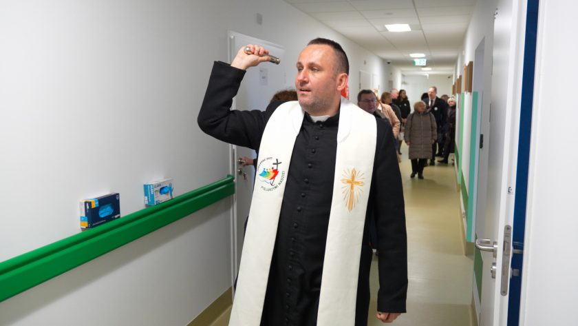 Kapłan w sutannie i białej stule idzie korytarzem nowej placówki, wykonując gest błogosławieństwa za pomocą metalowego kropidła. Za nim podąża grupa uczestników uroczystości. Korytarz jest jasny, z zielonymi poręczami i równomiernym oświetleniem