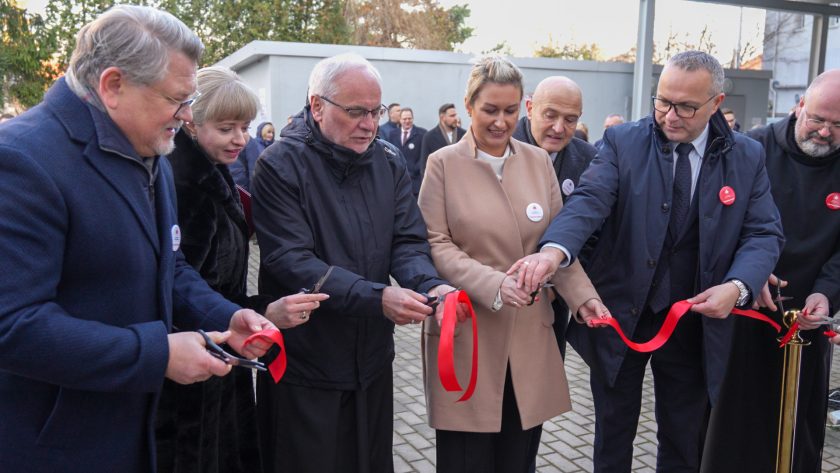 Grupa osób stoi obok siebie, trzymając nożyczki w rękach i wspólnie przecinając czerwoną wstęgę podczas ceremonii otwarcia. Uczestnicy uśmiechają się i patrzą na wstęgę. W tle widoczni są pozostali goście wydarzenia oraz zabudowania placówki