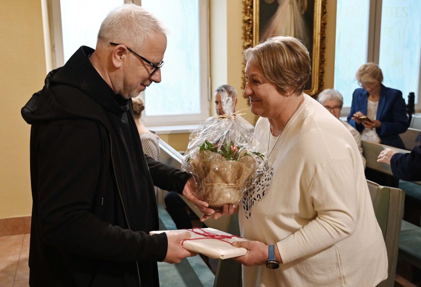 Zakonnik w czarnym habicie wręcza bukiet w celofanie kobiecie w białym swetrze, która uśmiecha się, trzymając także zapakowany prezent. W tle osoby siedzą w ławkach kaplicy