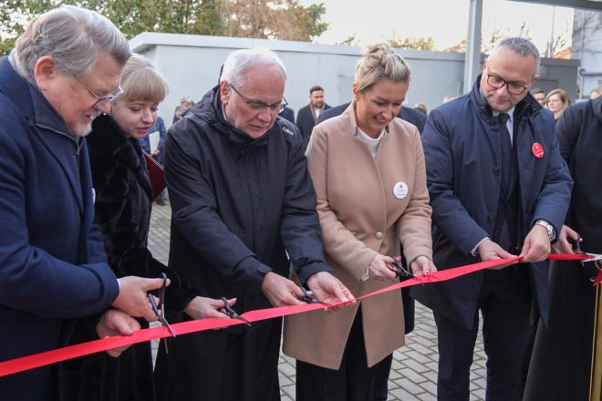 Kadr przedstawiający moment przecięcia czerwonej wstęgi. Każda osoba trzyma własne nożyczki. Ceremonię prowadzi pięć osób stojących w pierwszym rzędzie, ujętych z bliska – w tym osoba w beżowym płaszczu i duchowny w czarnym habicie