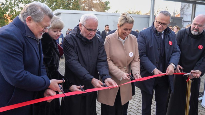 Kadr przedstawiający moment przecięcia czerwonej wstęgi. Każda osoba trzyma własne nożyczki. Ceremonię prowadzi pięć osób stojących w pierwszym rzędzie, ujętych z bliska – w tym osoba w beżowym płaszczu i duchowny w czarnym habicie