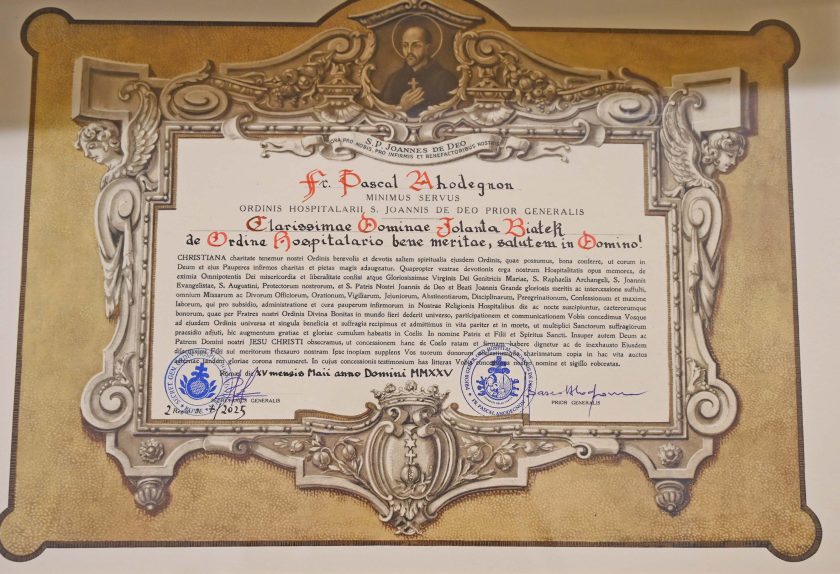 Zbliżenie na ozdobny, oprawiony w ramę dokument z ornamentami i wizerunkiem św. Jana Bożego u góry. Tekst sporządzony jest w języku łacińskim, a u dołu widoczne są pieczęcie i podpisy. Dokument dotyczy wyróżnienia osoby o imieniu Jolanta Biatek