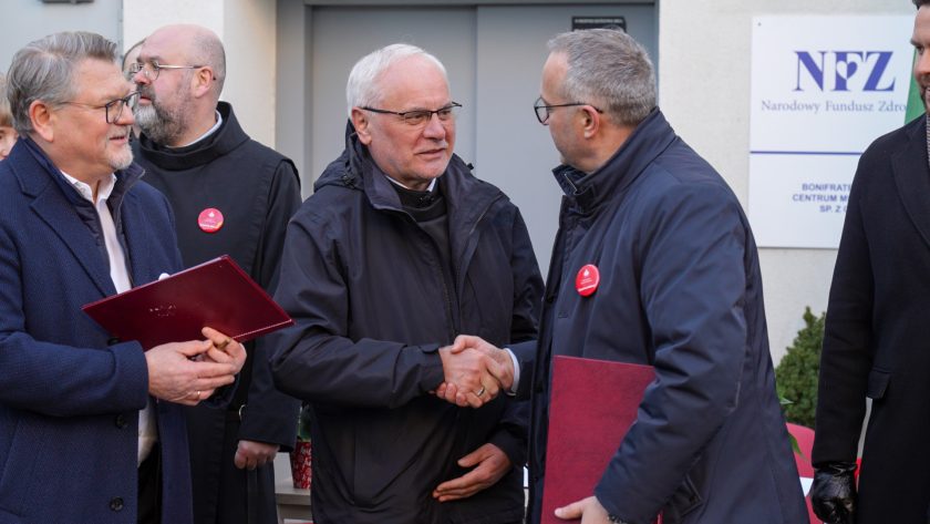 Trzech mężczyzn stoi przed wejściem do placówki. Dwóch z nich wymienia uścisk dłoni – jeden w ciemnej kurtce, drugi w granatowym płaszczu. Obaj trzymają bordowe teczki z dokumentami. W tle widoczne są osoby duchowne oraz tablica NFZ