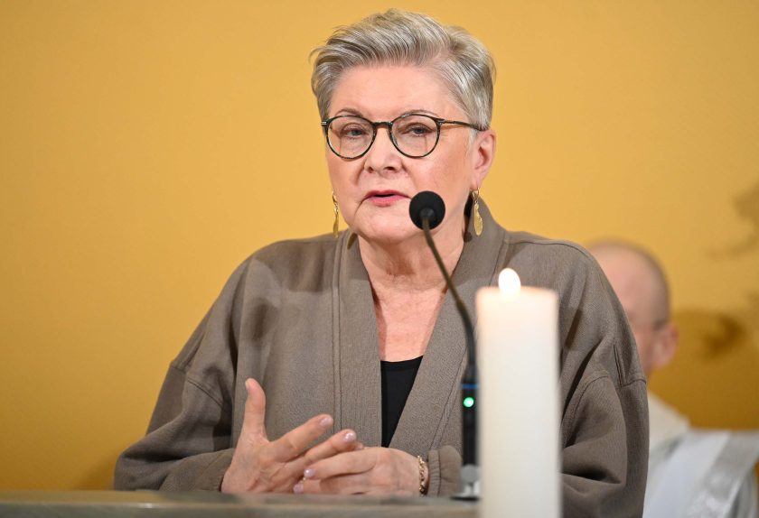 Kobieta z krótkimi siwymi włosami i okularami stoi przy ambonie, przemawiając do mikrofonu. Ma na sobie beżową marynarkę i długie kolczyki. Na pierwszym planie widoczna jest zapalona biała świeca, a w tle rozmyta sylwetka jednej z osób duchownych